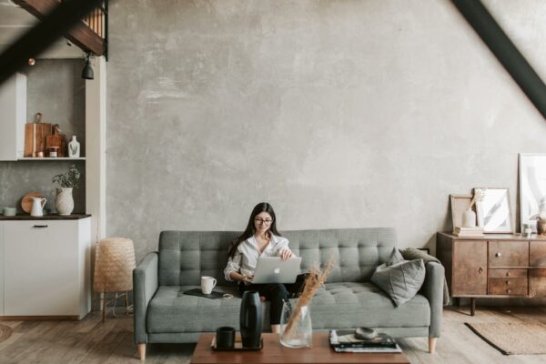 Happy female remote worker in eyeglasses sitting on cozy sofa with netbook and cup of coffee in loft style room with wooden floor and concrete wall